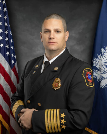 A uniformed firefighter stands in front of the US and South Carolina flags. He wears a dark double-breasted jacket with gold buttons, a patch on the shoulder, and gold-striped sleeves, and has a badge and insignia on the lapel.