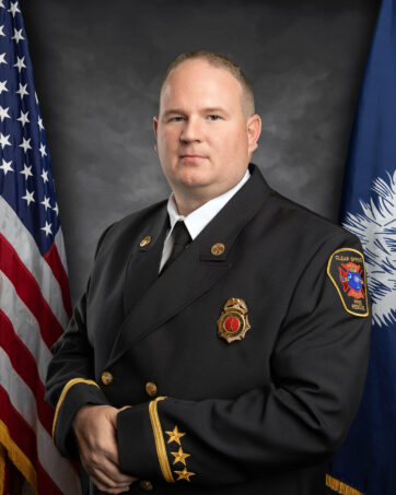 A firefighter in a formal uniform stands against a backdrop featuring the United States flag on one side and a blue flag on the other. He wears a badge and has gold stripes and stars on his sleeve.