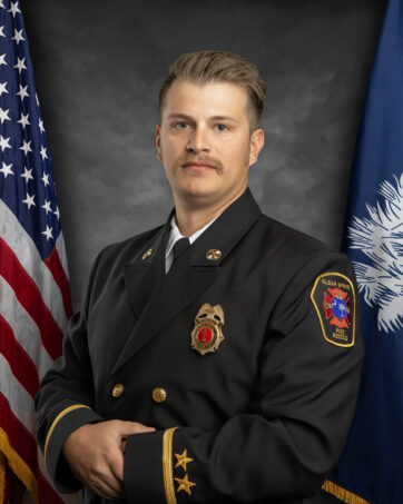 A firefighter in formal dress uniform stands between the U.S. flag and a blue flag with a white crescent and palmetto tree, against a gray background. He has a light brown mustache and short hair, with service insignias on his jacket.
