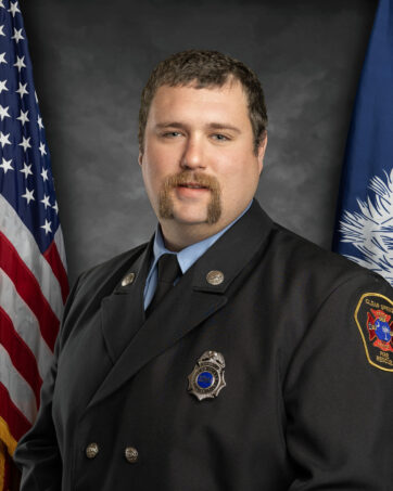 A person in a formal uniform poses for a portrait. The uniform includes a badge and insignia. They stand in front of the American flag and another flag with a palm and crescent design, against a gray background.