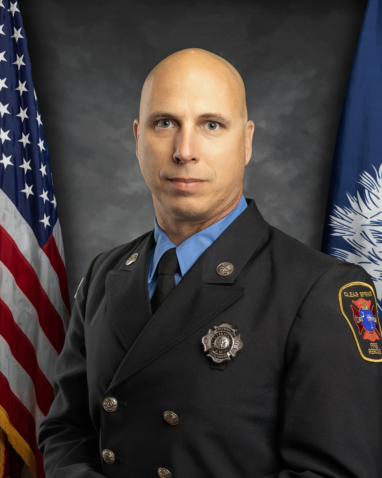 A person in a formal firefighter uniform poses for a portrait. They stand against a backdrop featuring the American flag and a blue flag with a white emblem. The uniform has badges and a patch on the shoulder.