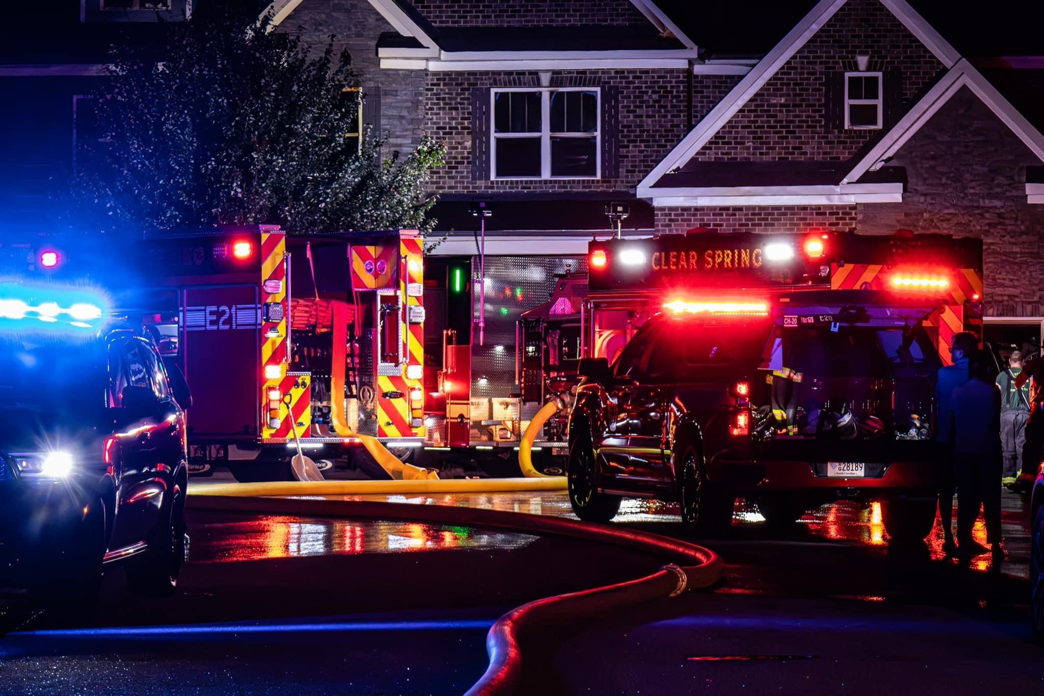 Fire trucks and emergency vehicles with flashing lights are parked in front of a residential building at night. Yellow hoses are spread across the wet road, and the area is dimly lit by the vehicles' lights.